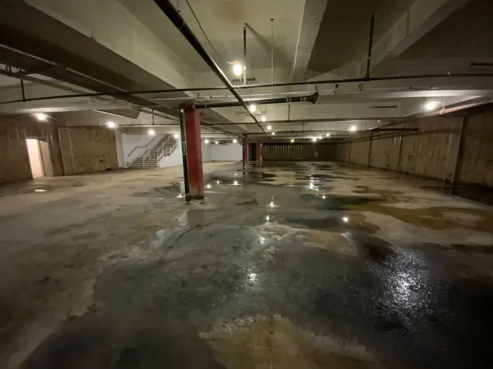 A large, dimly lit underground parking garage with wet, stained concrete floors suggests the need for mold investigation. The space appears vacant, with exposed pipes and a distant staircase leading to an exit. Fluorescent lights cast a faint glow across the area, emphasizing its eerie emptiness.