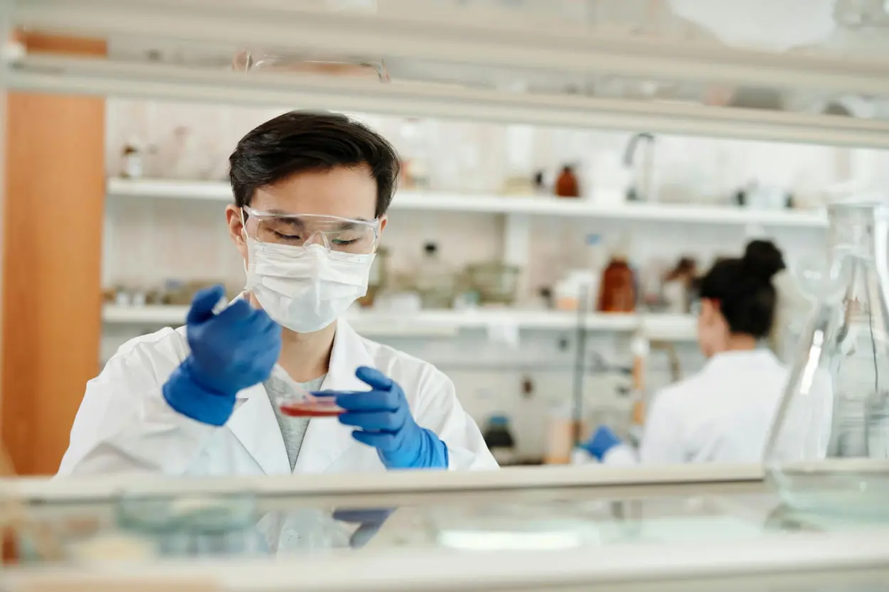A scientist, focused on testing, wears a mask, safety goggles, and gloves in the lab. They expertly use a pipette to transfer liquid into a petri dish. In the background, shelves brim with equipment and glassware, while another scientist conducts an inspection.