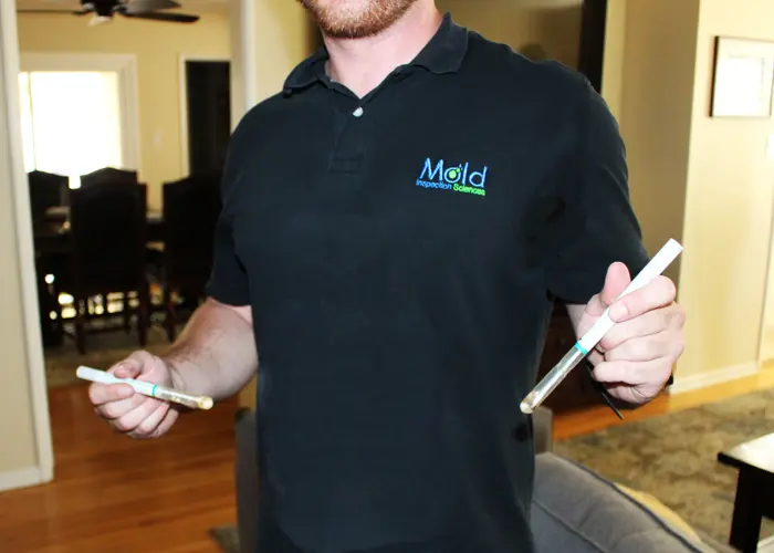 A person in a black shirt, emblazoned with "Mold Inspection Services," holds two sampling swabs in a home environment, likely conducting a mold investigation.