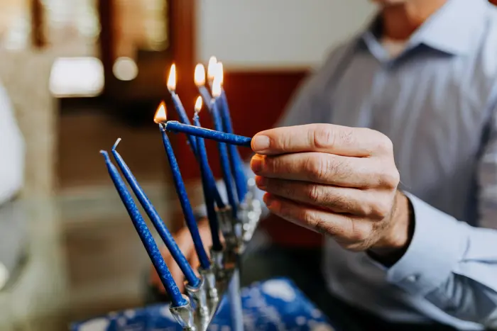 A person in a light blue shirt is carefully lighting blue candles on a menorah, conducting a quiet assessment of their placement. Several candles are already lit, casting a warm glow. The background is softly blurred.