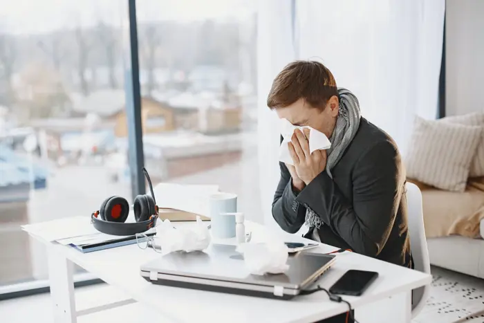 A person dressed in a suit and scarf sits at a desk, using a tissue to blow their nose. The desk has a laptop, headphones, a cup, and scattered tissues—perhaps an investigation into mold allergies. A large window offers a view of rooftops and trees outside.