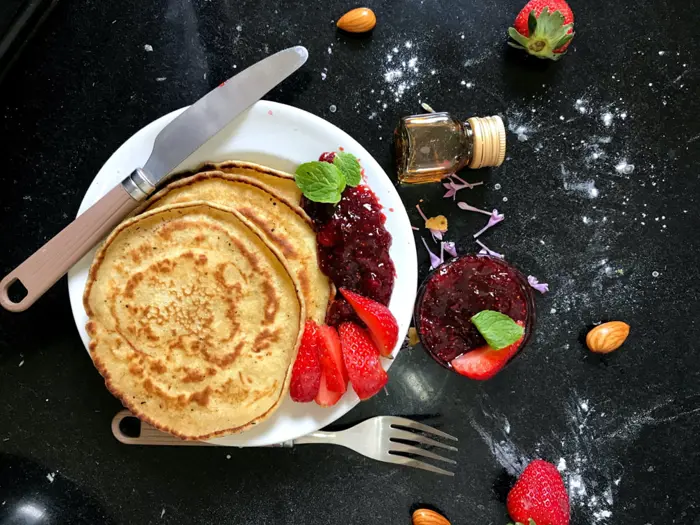 A plate of pancakes with sliced strawberries and red jam, garnished with mint leaves, awaits your taste test. A small bottle of syrup, whole almonds, and scattered flour surround the plate on a dark countertop. Nearby, a knife and fork are ready for your delicious assessment.