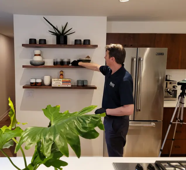 A person wearing a dark polo shirt and gloves carefully inspects items on wooden shelves in a modern kitchen. The space boasts a stainless steel refrigerator, a large leafy plant, and a tripod stand, creating an ideal setting for keen assessment.