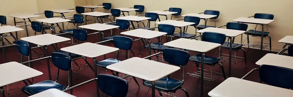 The empty classroom, primed for testing, features rows of blue chairs and white desks uniformly arranged on a red carpet. The light-colored walls enhance the bright and well-lit atmosphere.