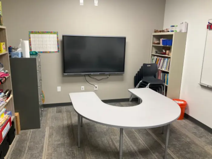 A classroom corner with a U-shaped table faces a large, wall-mounted flat-screen monitor. Shelves filled with books, stacked chairs, and a calendar are visible, alongside a trash can, various supplies for testing experiments, and even some mold samples.