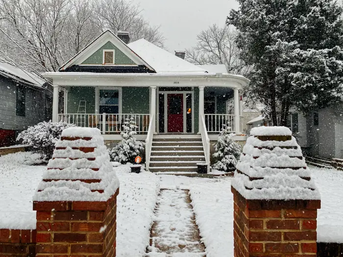 A cozy green house with a white porch is surrounded by snow-covered trees and a yard. Snowflakes fall gently, blanketing the roof and steps. Brick pillars frame the walkway leading to the front door, creating a picturesque winter scene perfect for testing your camera skills.