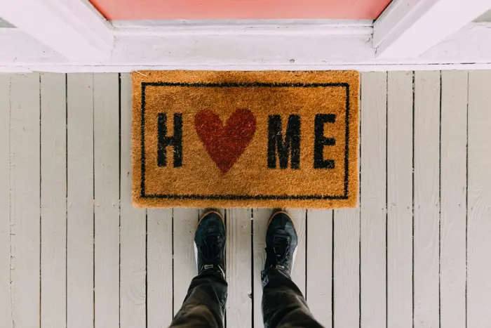 A person stands in front of a doormat that reads HOME with a red heart symbol replacing the O, placed on a light gray wooden porch. They pause for an inspection of their surroundings, wearing black shoes, as they savor the feeling of being welcomed back.