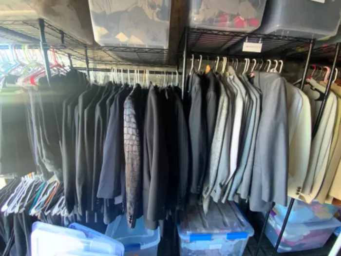 A storage room with metal shelves holding meticulously organized rows of clothing items on hangers, primarily suits in various shades of black and grey, stands ready for assessment. Clear plastic bins with lids are stacked above and below the hanging clothes.